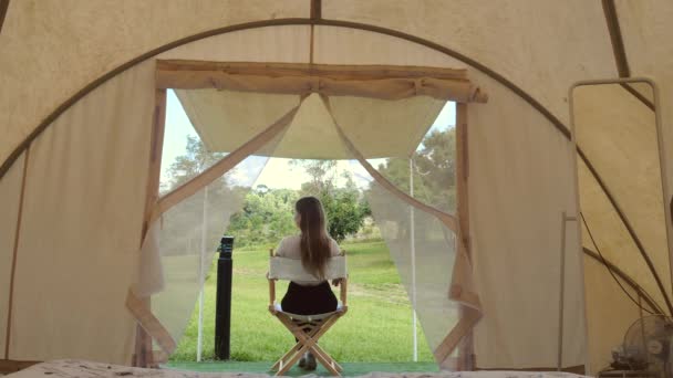 Viaggio campeggio donna seduta all'esterno della tenda, relax e vista sulla natura e gli animali selvatici. Vista posteriore di turista femminile in vacanza in tenda moderna. Disintossicazione digitale. Concetto glamping, eco turismo. — Video Stock