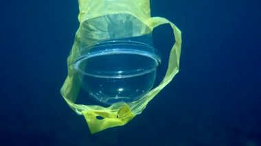 Plastik bardakla kristal mavi suda güneş ışınlarında yüzen plastik bir bardağın sualtı videosu. Çevresel plastik kirliliği sorunları kavramı. Dünya okyanus günü. Okyanustaki çöp..
