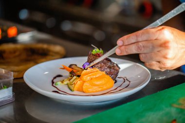 Otel ya da restoran mutfak dekorasyon tabağında aşçı yamaklarını kapatın. Bifteği patates püresiyle hazırlamak, sebze eklemek, özel cımbızla dekore etmek. Şefin elleri et pişiriyor.