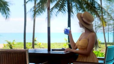 Tropik tatil beldesinde yaz tatili. Sahildeki kafede oturan ve içecek bir şeyler içen kadın deniz ve palmiye ağaçlarının tadını çıkarıyor..