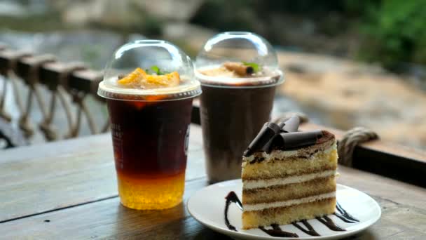 冷たい飲み物とチョコレートケーキの一部が付いている2つのカップは 滝を背景に屋外カフェで木製のテーブルに立っています — ストック動画