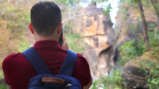携帯電話で峡谷を撮影した森の中の30代の山岳ハイキング写真家やビデオブロガーの男 赤いポロの男旅行者とバックパックのビデオカメラマンが自然の中に立っている バックビュー — ストック動画