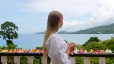 Romantik bir tatilde beyaz otel bornozuyla bir fincan sıcak sabah kahvesi içen çekici genç bir kadın. Modern tropikal tatil köyünde okyanus manzaralı kadın gezginin yan görüntüsü. Seyahat yaşam tarzı