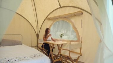 Kamp çadırındaki kadın, pencerenin yanında oturuyor, doğanın ve içme suyunun tadını çıkarıyor. Yaz sabahı ormanda kamp yaparken. Aktif yaşam tarzı, seyahat, tatil, kamp, eko turizm konsepti