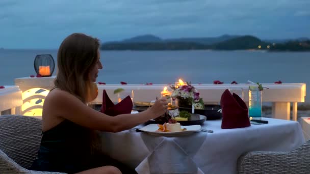 エレガントな夜のガウンを着た幸せな女性は 高級ホテルのレストランで夜にキャンドルとロマンチックなディナーを持って おいしい食べ物を食べる 海を背景に 熱帯休暇や休日の女性観光客 — ストック動画