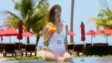 Yüzme havuzunun kenarında oturan, elinde kokteyl ile güneşlenen genç ve mutlu bir kadın. Şapkalı gülümseyen kız, tatillerde tropik otellerde ya da tatil köylerinde dinleniyor. Yaz tatili.