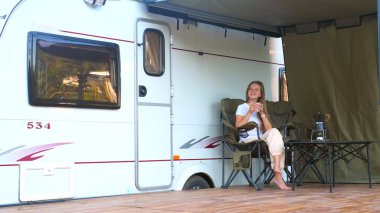 Kadın elinde bir kupa kahveyle karavanın yanında oturuyor, doğanın tadını çıkarıyor. Karavan tatili. Aile seyahati, karavanla tatil gezisi.