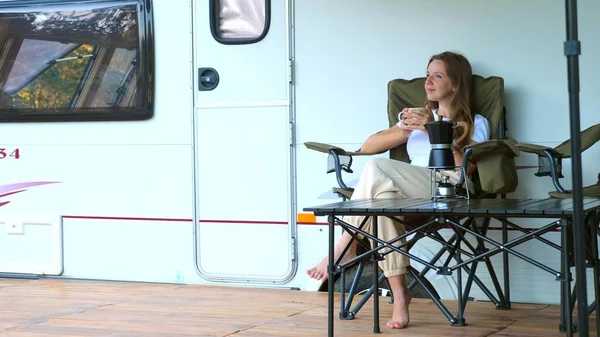 Smiling woman sitting on folding chair, enjoy evening and sunset on camp site next to camper RV. Concept family vacation, holidays and summer