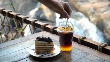 Erkek eli soğuk turuncu kahve ile plastik bardağa bir tüp yerleştirir. Buzlu kahve, doğa geçmişi olan bir kafeye gidecek. Garson siparişi içecek ve tatlı pastayla servis ediyor.