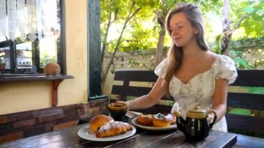 Açık kafede sıcak çay içen bir kadın, tabaklar dolusu taze hamur işiyle masada oturuyor, fırının ve arka planda yeşil ağaçların tadını çıkarıyor..