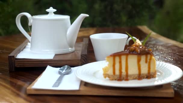 緑豊かな自然を背景に テーブルの上に急須で熱いお茶やセラミックティーカップを蒸しクローズアップ 屋外カフェでのホットドリンクとキャラメルケーキ — ストック動画