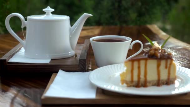 急須でテーブルの上のセラミックティーカップにスプーンで熱々のお茶をかき混ぜて 自然を背景にしたソーサーに甘いケーキを入れます 屋外カフェでのキャラメルソースチーズケーキ — ストック動画