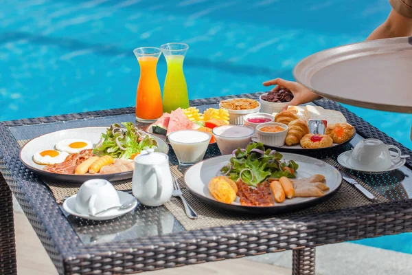 Waiter hand serving breakfast in restaurant at luxury hotel. Asian woman waitress with tray put morning food, drinks on table in outdoor pool cafe in tropical resort. Travel, holiday, vacation concept