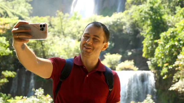 Happy social media content maker. Smiling influencer talk on video, shoot blog on mobile phone outdoors. Adult man blogger, vlogger recording selfie movie on his smartphone at nature, look at camera.