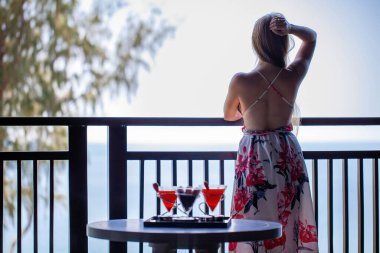 Modern tropikal otelin deniz manzaralı balkonunda duran zarif elbiseli etkileyici kadının arka görünüşü. Plajda egzotik yaz tatilindeki gezgin kız. 