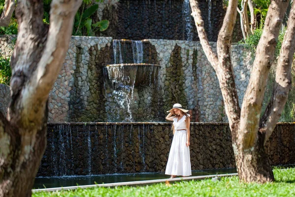 stock image Travel woman in hat walking near waterfall in modern tropical resort. Female tourist in white dress relaxing on beautiful hotel territory. Concept of summer vacation and weekend