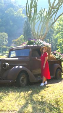 Klasik klasik arabanın yanında yürüyen kırmızı elbiseli ve şapkalı şık bir kadın. Arka planda yaşlı bir kamyonet ve kaktüsün yanında parkta yürüyen çekici, modern bir kadın. Dikey video