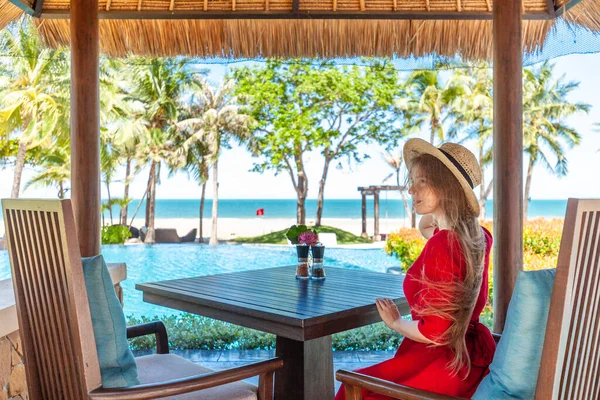 Woman on exotic summer vacation. Travel girl in red dress and hat sitting on breakfast at the table in hotel lounge near pool. Tropical beach restaurant with sea view