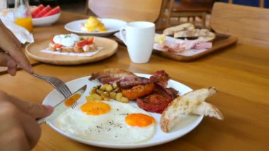 Adam kafede kızarmış ekmek, pastırma ve domatesle kızarmış yumurta yiyor. Kahvaltı ya da öğle yemeği. Çatal ve bıçaklı erkek eller, güneşli kısımları, yumurtaları, beyaz tabakta, restoranda beyaz tabakta kesiyorlar..