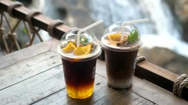 Paket servis için pipetli plastik bardaklarda içecek. Arka planda şelale olan açık kafede buzlu portakallı kahve ve çikolatalı kakao içecekler. Doğa Kafesi