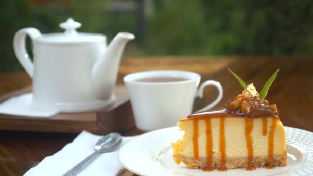 在户外咖啡店的木制桌子上 放着一块美味的新鲜芝士蛋糕和一个白色茶壶 用可食用的黄金和焦糖装饰的美味新鲜糕点和一杯热茶 英语茶点时间 — 图库视频影像