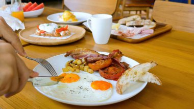 Adam kafede kızarmış ekmek, pastırma ve domatesle kızarmış yumurta yiyor. Kahvaltı ya da öğle yemeği. Çatal ve bıçaklı erkek eller, güneşli kısımları, yumurtaları, beyaz tabakta, restoranda beyaz tabakta kesiyorlar..