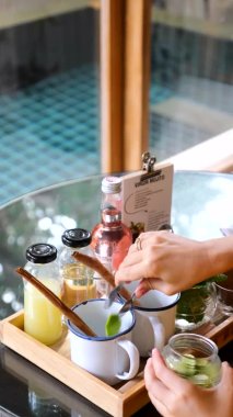 Bakire mojito hazırlarken çekilmiş dikey bir video. Kadın elleri, tarçın çubuğuyla limon dilimlerini bardağa koyar. Arka planda havuz manzaralı tropikal bir villada içecek hazırlıyorum.