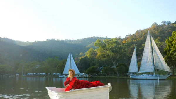 Romantic travel vacation. Stylish woman in red dress on date, sitting in decorated boat with flowers, enjoy sunset on the lake. Cute elegant girl celebrate birthday or anniversary