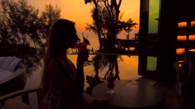 Gün batımında tropik oteldeki lüks restoranda meyve alkollü kokteyl içen genç bir kadının silüeti. Akşam üstü meyhane barında elinde kokteyl tutan kadın turist. Güzel turuncu gökyüzü.