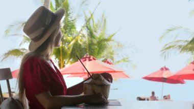 Tropik plaj tatil beldesinde yaz tatili. Şapkalı, açık kafede oturan, taze hindistan cevizi içen ve deniz manzarasının tadını çıkaran mutlu bir seyahat kadını. Tatilde olan kadın gezgin.. 