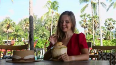 Yaz tatili konsepti. Mutlu gezgin kadın, Tayland 'ın Khao Lak plajındaki taze hindistan ceviziyle kafede dinleniyor. Arka planda palmiye ağaçları olan açık hava restoranında dinlenen neşeli kız.