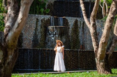 Modern tropikal tatil köyünde şelalenin yanında yürüyen şapkalı bir kadın. Beyaz elbiseli kadın turist güzel otel bölgesinde dinleniyor. Yaz tatili ve hafta sonu kavramı