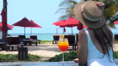 Yüzme havuzunda hasır şapka ve mayo giymiş genç ve güzel bir kadın, sıcak kırsalda tatili bir bardak turuncu kokteyl ile kutluyor. Tropik yaz tatili