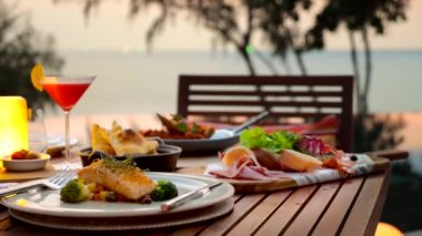 Günbatımında ahşap masada romantik bir akşam yemeği sunuldu. Deniz, ufuk, gökyüzü arka planı. Lüks otel ya da tatil köyünde güzel bir restoran. Seramik tabakta brokolili ızgara somon, kokteyl, soğuk et..