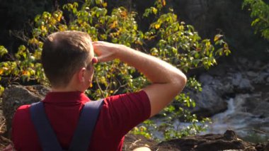 Kanyon kenarındaki sırt çantalı adam alnında bir şey ararken ufka bakıyor. Erkek gezgin uzaklara bakıyor, doğanın, gün batımının ve nehir manzarasının tadını çıkarıyor.