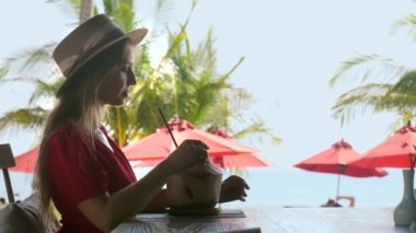 Tropik tatil. Arka planda palmiye ağaçları ve deniz olan hindistan cevizi suyu içen genç bir kadın. Yaz tatilinde sahil kafede dinlenmek. Bayan gezgin Tayland tatilinin tadını çıkarıyor.