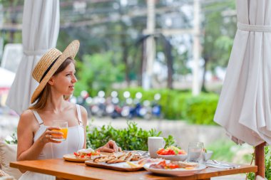 Oteldeki açık hava restoranında kahvaltıda gülümseyen genç bir kadın. Hasır şapkalı kadın gezgin modern kafede portakal suyu içiyor. Yaz tatili