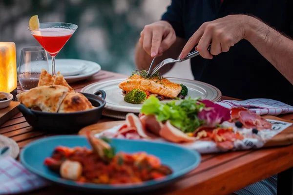 Man on luxury dinner served on table with fresh seafood, cold cuts and cocktail glass in luxury outdoor restaurant. Man hands cutting grilled salmon. Healthy food