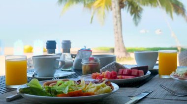 Tropikal otelde sabah yemeği. Sağlıklı vejetaryen yiyeceklerle dolu bir masa. Meyve, meyve suyu, deniz kenarındaki tatil köyünde salata ve arka planda hurma ağaçları. Yaz tatili, Tayland tatili konsepti