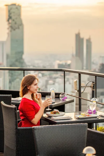 Beautiful young woman with cocktail rests at luxury rooftop restaurant at sunset. Elegant female in red dress with drink at sky bar terrace looking at modern city skyline. Skyscrapers on background.