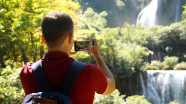 Seyahat halinde sırt çantalı mutlu bir erkek turist. Seyahat tutkusu, turizm konsepti. Genç adam gezgin, akıllı telefon kamerasında sosyal medya için inanılmaz şelaleler ve tropikal doğa videoları yapıyor..