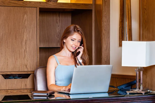 Young smiling business woman using laptop computer, calling by phone in modern apartment. Attractive businesswoman working from luxury hotel in business trip, calls with clients.
