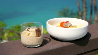 Healthy breakfast with oatmeal and fruits yogurt salad. Assorted fresh tropical chopped fruits in bowl. Oatmeal porridge with nuts, almonds and raisins with sea on background.