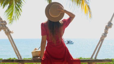 Şapkalı ve kırmızı elbiseli turist genç hindistancevizli ahşap salıncakta oturuyor ve deniz ve palmiye manzarasının tadını çıkarıyor. Tropik tatil beldesinde yaz tatili. Sahildeki otel, arka planda yat.
