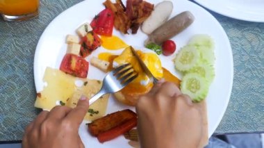 Kadın eli Benedict yumurtasını çatal ve bıçakla Hollandaise sosuyla kesiyor. Haşlanmış yumurta, pastırma, sosis, peynir, açık büfede ızgara sebze kahvaltı, otelde restoranda brunch, tatil köyü..