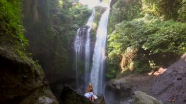 Young travel woman sitting on the rock in front of Aling Aling waterfall among tropical rainforest and plants on the north of Bali island, Indonesia. Concept of adventure, Asian trip and vacation