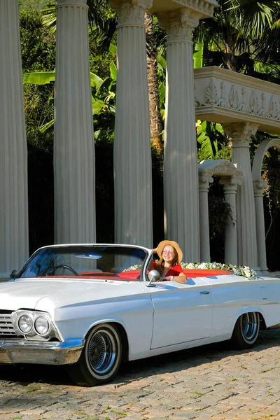 Travel woman in red dress and straw hat sitting in convertible vintage retro car next to the antique columns and enjoy tropical summer day. Concept of fashion, style and beauty
