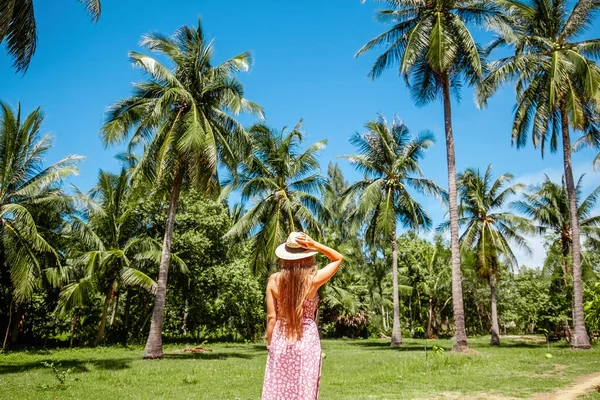 Back view of travel women over exotic tropical green palm trees and blue sky background. Summer vacation. Girl in straw hat relaxing on travel vacation in summer sun on Thailand island.