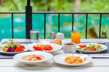 Kahvaltı, lüks bir tropikal otel veya tatil köyündeki açık hava restoranında servis edilir. Yemek ve içeceklerle birlikte kahvaltı masası. Yaz tatilleri, tatiller, turizm, seyahat konsepti. Yeşil gölet arka planı.