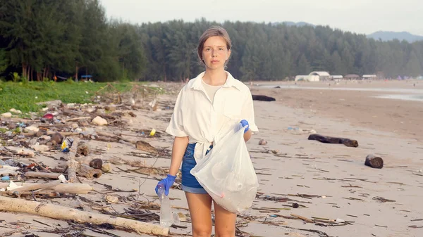 Young woman volunteer shows the camera where to put trash in nature, collecting trash in a trash bag on the sea beach. Plastic ocean pollution and environmental problem, ecosystem concept.